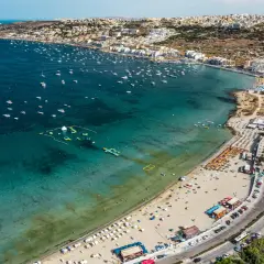 Malta zachwyca turkusowym morzem pełnym łodzi i wodnych atrakcji. Plaże tętnią życiem, a miasto rozciąga się wzdłuż wybrzeża.