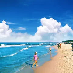 Słoneczna plaża, fale, dzieci bawiące się w piasku i w wodzie. W tle widać las i ludzi cieszących się wakacjami. Idealne miejsce na chałupy imprezy.