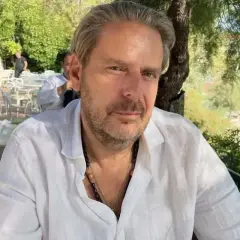 Mężczyzna w białej koszuli, z naszyjnikami, siedzi przy stoliku w restauracji. W tle widać plażę i drzewa. To może być jego atelier amaro.