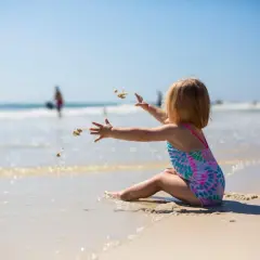 Dziewczynka w kolorowym stroju kąpielowym rzuca piasek na plaży. Idealny obrazek do poradnika: co zabrać na wakacje nad morze z dzieckiem.