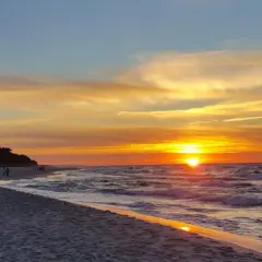 Zachód słońca w Jastarni maluje niebo odcieniami złota i pomarańczy. Fale delikatnie szumią na piaszczystej plaży, a sylwetki ludzi spacerują brzegiem.