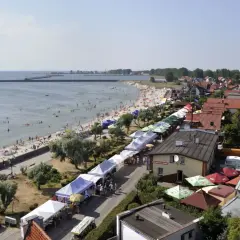 Widok z góry na plażę z ludźmi, deptak z kramami i zabudowania nad morzem.