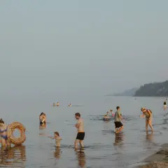 Ludzie kąpią się i spacerują po plaży, podczas gdy nad morzem unosi się delikatna mgła. W oddali widać statek.