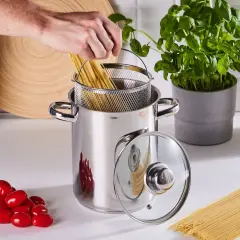 Ręka wkłada spaghetti do garnka, rozpoczynając gotowanie makaronu. Obok pomidorki i świeża bazylia.