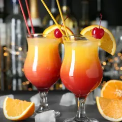 Dwa drinki w domu, ozdobione pomarańczą i wiśnią, z kostkami lodu i owocami w tle.