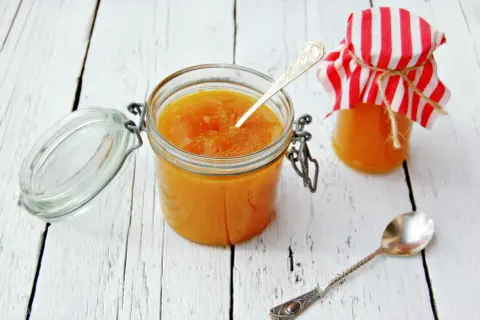 Słoiki z domowym dżemem z mirabelek, jeden otwarty z łyżeczką, drugi ozdobiony czerwono-białą chusteczką.