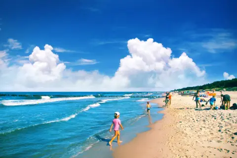 Słoneczna plaża, fale, dzieci bawiące się w piasku i w wodzie. W tle widać las i ludzi cieszących się wakacjami. Idealne miejsce na chałupy imprezy.