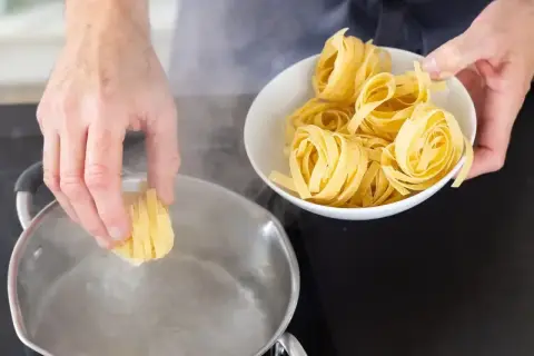 Ręka wkłada porcję tagliatelle do wrzącej wody. Czy makaron gotujemy pod przykryciem? Zobacz, jak zacząć gotowanie.
