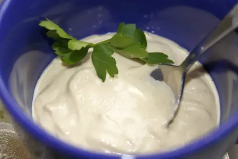 Kremowy sos z tahini, udekorowany gałązką pietruszki, czeka na łyżce w niebieskiej miseczce.