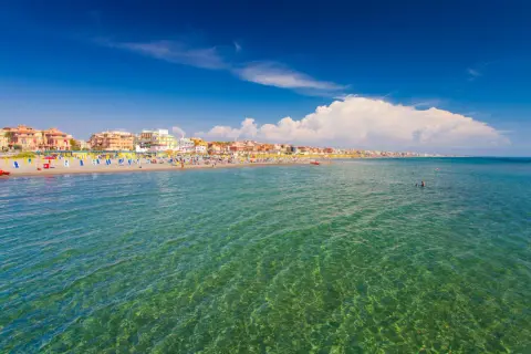 Słoneczny dzień nad morzem w Rzymie. Tłumy ludzi na plaży, kolorowe parasole i budynki w tle.