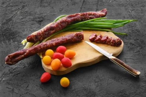 Dwa długie kabanosy, pokrojone plastry, pomidorki i szczypiorek na desce. Czy kabanosy są zdrowe? To pytanie zadaje sobie wielu.
