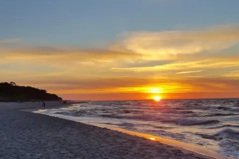 Zachód słońca w Jastarni maluje niebo odcieniami złota i pomarańczy. Fale delikatnie szumią na piaszczystej plaży, a sylwetki ludzi spacerują brzegiem.