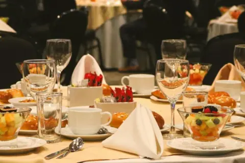 Elegancko nakryty stół w hotelowym lobby, gotowy na śniadanie. Widać owoce, pieczywo, filiżanki i kieliszki.