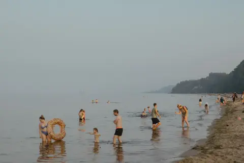 Ludzie kąpią się i spacerują po plaży, podczas gdy nad morzem unosi się delikatna mgła. W oddali widać statek.