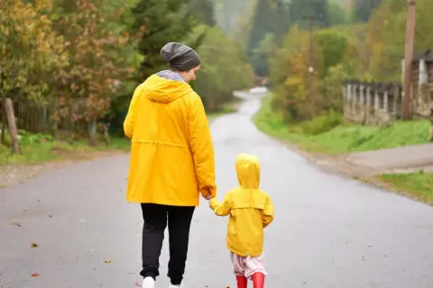 Mama i dziecko w żółtych płaszczach idą za rękę mokrą drogą. Nawet w deszczowe dni Jastarnia oferuje atrakcje, jak spacery po malowniczych okolicach.