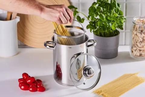 Ręka wkłada spaghetti do garnka, rozpoczynając gotowanie makaronu. Obok pomidorki i świeża bazylia.