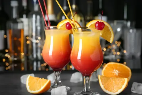 Dwa drinki w domu, ozdobione pomarańczą i wiśnią, z kostkami lodu i owocami w tle.