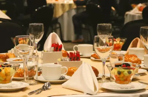 Elegancko nakryty stół w hotelowym lobby, gotowy na śniadanie. Widać owoce, pieczywo, filiżanki i kieliszki.