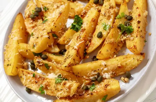 Ziemniaki po grecku, pieczone ćwiartki ziemniaków z ziołami, kaparami i parmezanem, wyglądają apetycznie.