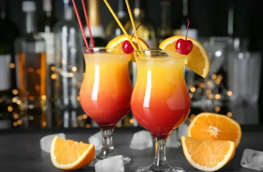 Dwa drinki w domu, ozdobione pomarańczą i wiśnią, z kostkami lodu i owocami w tle.