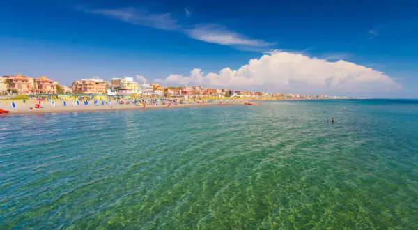 Słoneczny dzień nad morzem w Rzymie. Tłumy ludzi na plaży, kolorowe parasole i budynki w tle.