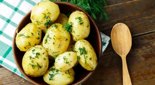 Miska pełna aromatycznych, gotowanych ziemniaków posypanych świeżym koperkiem, obok drewniana łyżka.