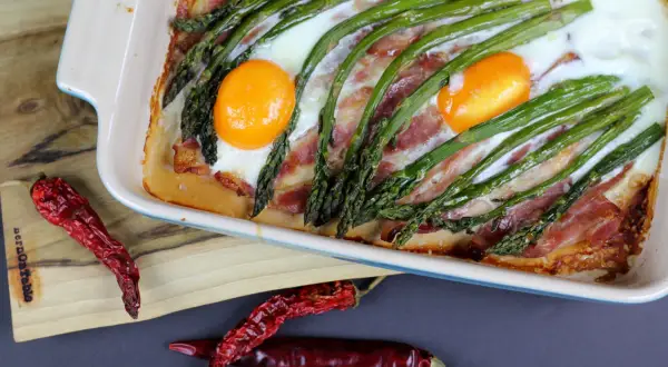 Pyszne szparagi na śniadanie z boczkiem i jajkami zapieczone w naczyniu. Obok leżą suszone papryczki chili.