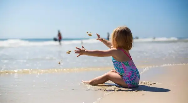 Dziewczynka w kolorowym stroju kąpielowym rzuca piasek na plaży. Idealny obrazek do poradnika: co zabrać na wakacje nad morze z dzieckiem.
