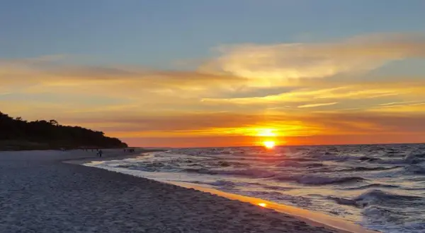 Zachód słońca w Jastarni maluje niebo odcieniami złota i pomarańczy. Fale delikatnie szumią na piaszczystej plaży, a sylwetki ludzi spacerują brzegiem.