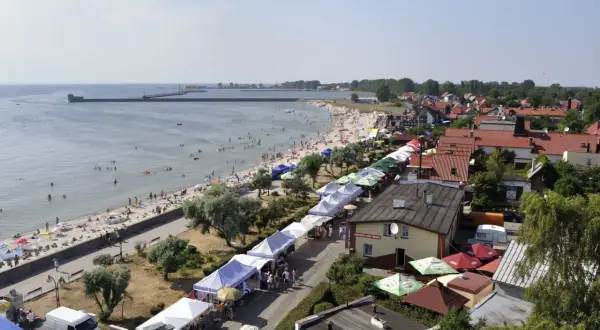 Widok z góry na plażę z ludźmi, deptak z kramami i zabudowania nad morzem.