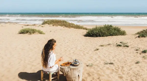 Kobieta pije herbatę na piaszczystej plaży. W tle widać turkusowe morze, które przypomina o pięknie Tunezji.