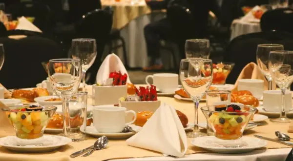 Elegancko nakryty stół w hotelowym lobby, gotowy na śniadanie. Widać owoce, pieczywo, filiżanki i kieliszki.