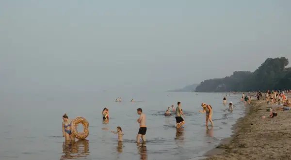 Ludzie kąpią się i spacerują po plaży, podczas gdy nad morzem unosi się delikatna mgła. W oddali widać statek.