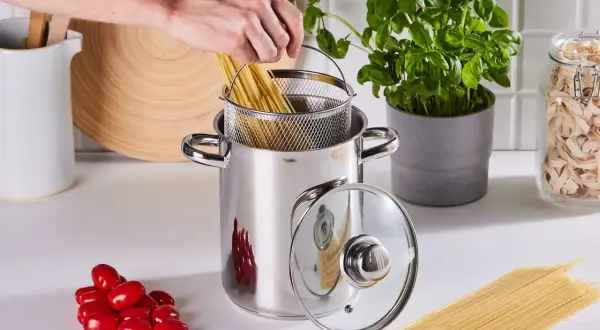 Ręka wkłada spaghetti do garnka, rozpoczynając gotowanie makaronu. Obok pomidorki i świeża bazylia.