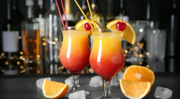 Dwa drinki w domu, ozdobione pomarańczą i wiśnią, z kostkami lodu i owocami w tle.