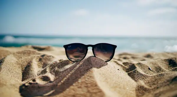 Okulary przeciwsłoneczne na piaszczystej plaży, w tle morze i błękitne niebo. Idealne na wakacje, gdy obowiązuje opłata klimatyczna nad morzem.