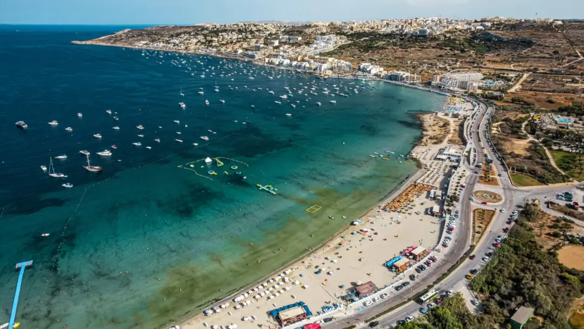 Malta zachwyca turkusowym morzem pełnym łodzi i wodnych atrakcji. Plaże tętnią życiem, a miasto rozciąga się wzdłuż wybrzeża.