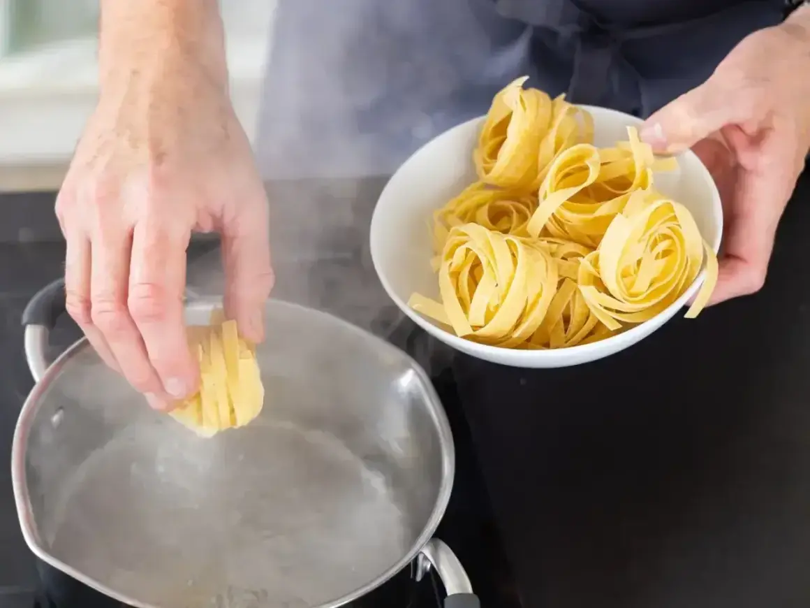 Ręka wkłada porcję tagliatelle do wrzącej wody. Czy makaron gotujemy pod przykryciem? Zobacz, jak zacząć gotowanie.