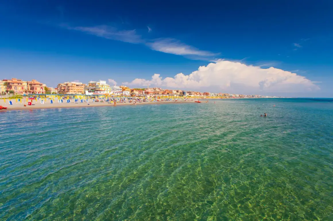 Słoneczny dzień nad morzem w Rzymie. Tłumy ludzi na plaży, kolorowe parasole i budynki w tle.