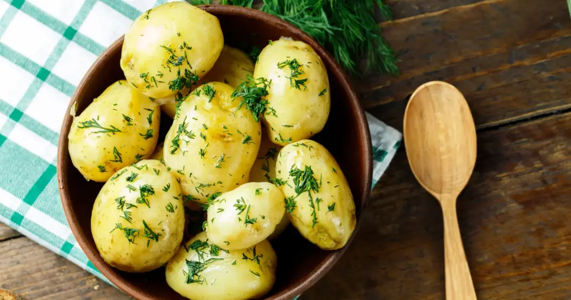 Miska pełna aromatycznych, gotowanych ziemniaków posypanych świeżym koperkiem, obok drewniana łyżka.