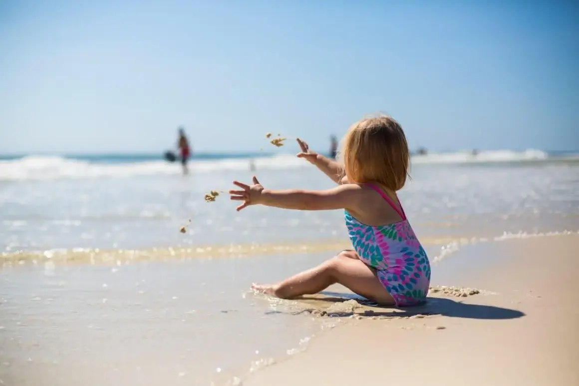 Dziewczynka w kolorowym stroju kąpielowym rzuca piasek na plaży. Idealny obrazek do poradnika: co zabrać na wakacje nad morze z dzieckiem.