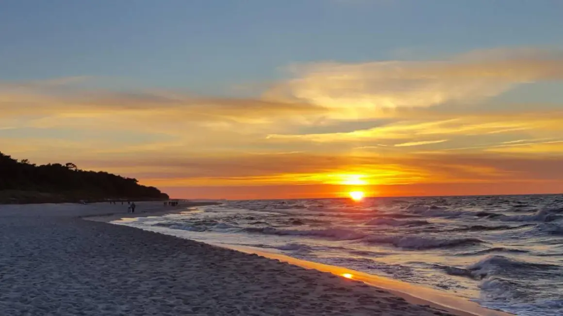Zachód słońca w Jastarni maluje niebo odcieniami złota i pomarańczy. Fale delikatnie szumią na piaszczystej plaży, a sylwetki ludzi spacerują brzegiem.