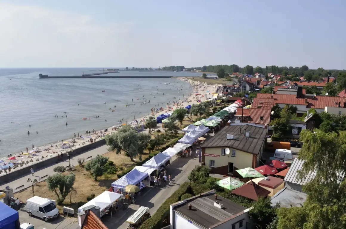 Widok z góry na plażę z ludźmi, deptak z kramami i zabudowania nad morzem.