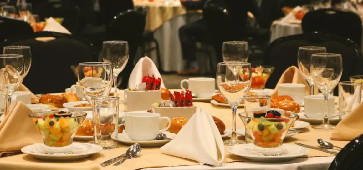 Elegancko nakryty stół w hotelowym lobby, gotowy na śniadanie. Widać owoce, pieczywo, filiżanki i kieliszki.