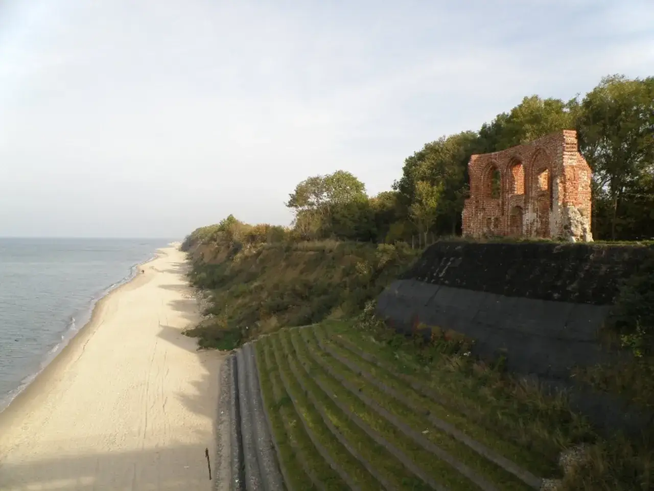 Ruiny ceglanego budynku na klifie nad piaszczystą plażą. Linia brzegowa Bałtyku z zielonymi tarasami.