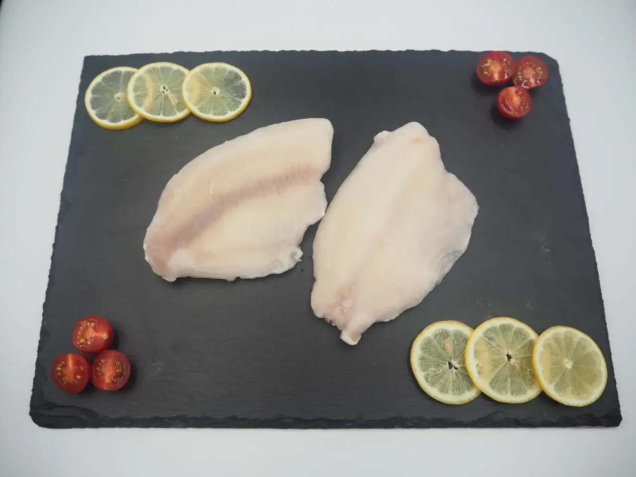 Dwa filety rybne, prawdopodobnie miruna lub tilapia, ułożone na czarnej desce z plasterkami cytryny i pomidorkami koktajlowymi.