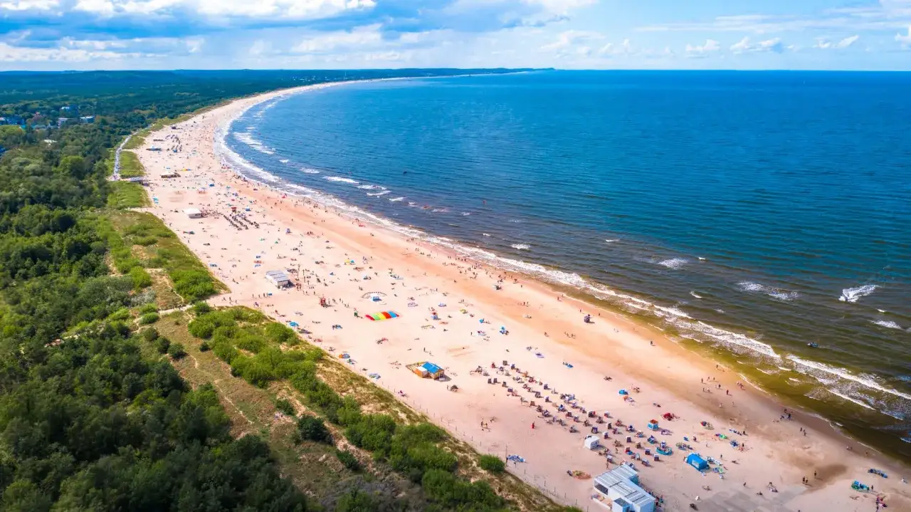 Tłum ludzi wypoczywa na szerokiej, piaszczystej plaży nad błękitnym morzem. Idealne miejsce, by zapytać: gdzie nad morze?
