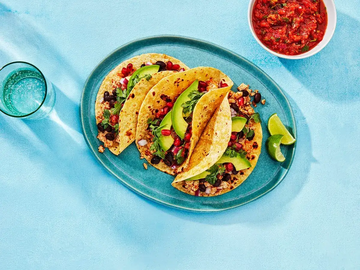 Pyszne tacos z awokado na śniadanie, z czarną fasolą, kolendrą i limonką. Obok miseczka salsy i szklanka wody.