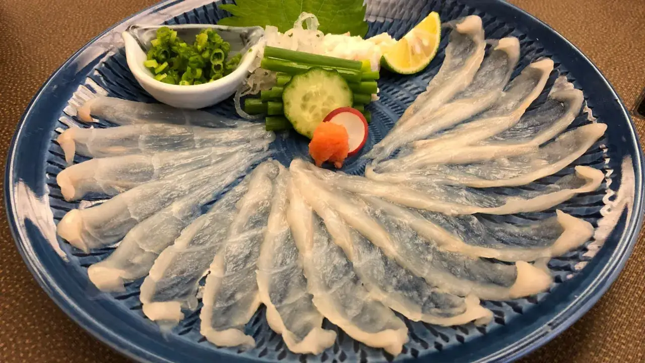Wykwintne sashimi z ryby fugu ułożone wachlarzowo na niebieskim talerzu, z dodatkami: szczypiorkiem, ogórkiem, rzodkiewką i cytryną.