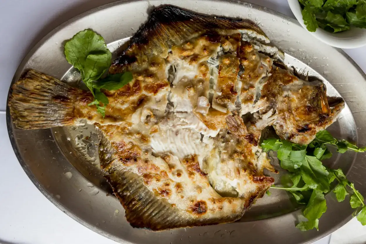 Złocisty, pieczony turbot na metalowej tacy, otoczony zielonymi liśćmi. Ten turbot smak kusi apetycznym wyglądem.